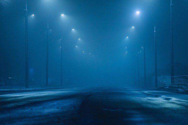 Dark foggy roadway at night barely illuminated by street lights.