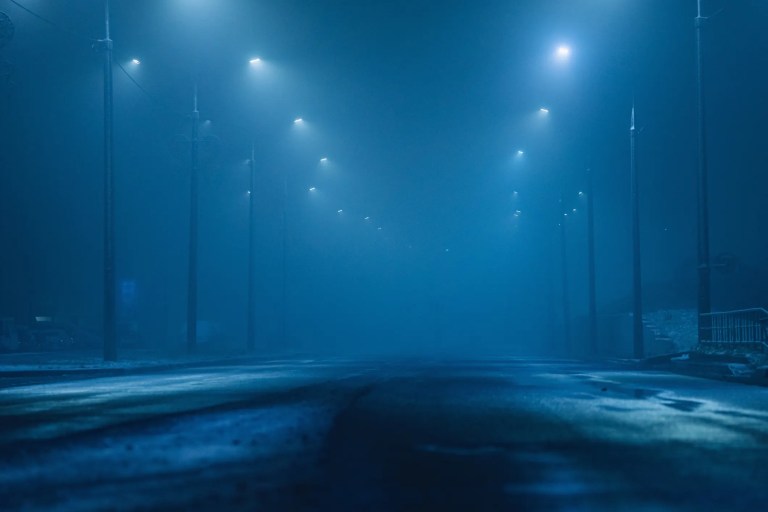 Dark foggy roadway at night barely illuminated by street lights.