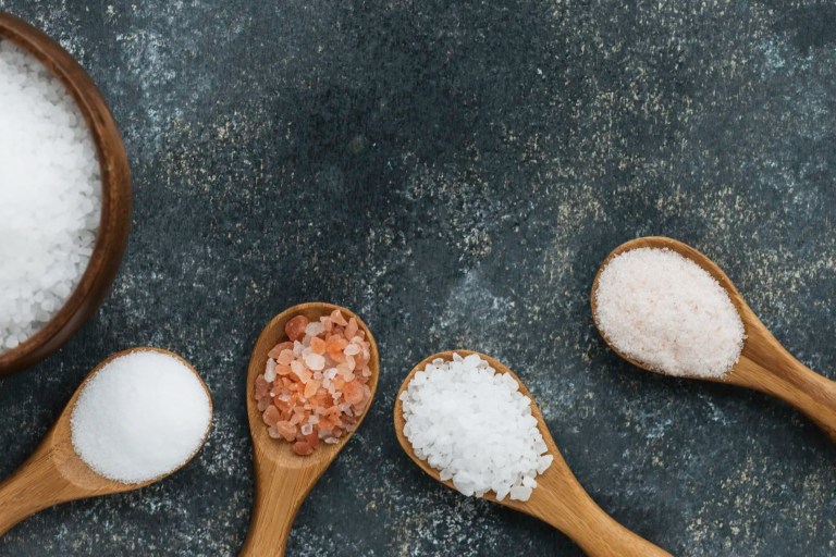 Four different types of salt in wooden spoons beside bowl of salt on stone background