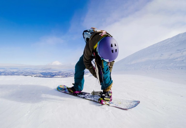 Snowboarder strapping in wearing purple helmet at top of slope