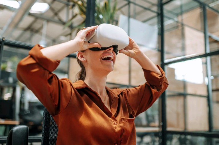 Woman in orange blouse wearing white VR headset and smiling in glass office