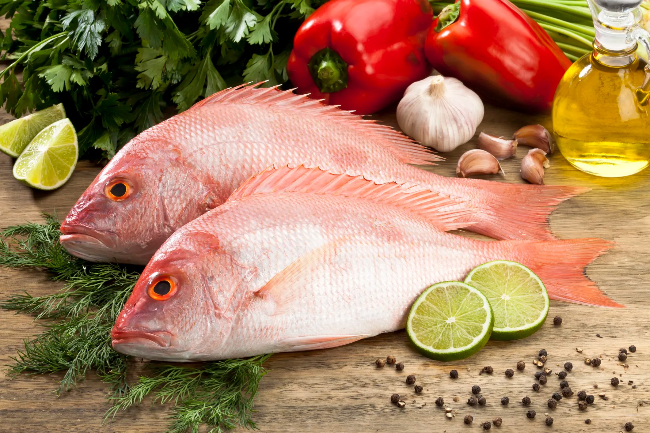 Two red snapper on board with lime slices, red peppers, olive oil, and herbs