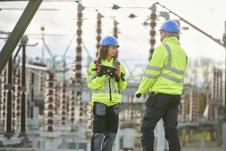 Two substation power workers wearing HVSA and ANSI Z87.1 blue hardhats with power lines in background.