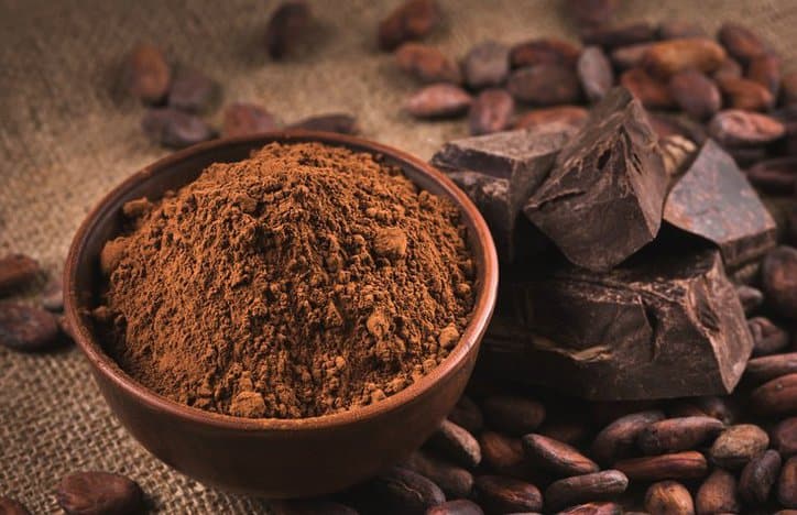 A bowl of cocoa powder next to pieces of chocolate and cocoa beans