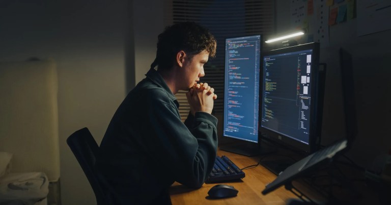 Young man working late coding on two monitors at desk in dark room to set the standard for lossless data compression.