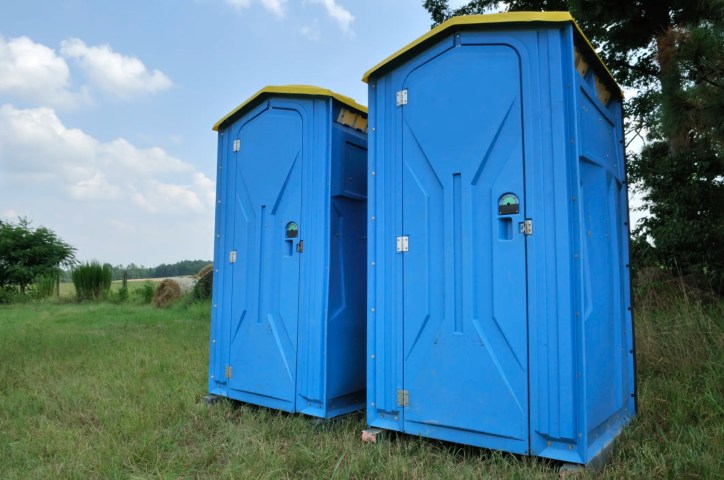 Two blue porta potties set up in grass to follow ANSI/ASSP A10.25 sanitation reqs.