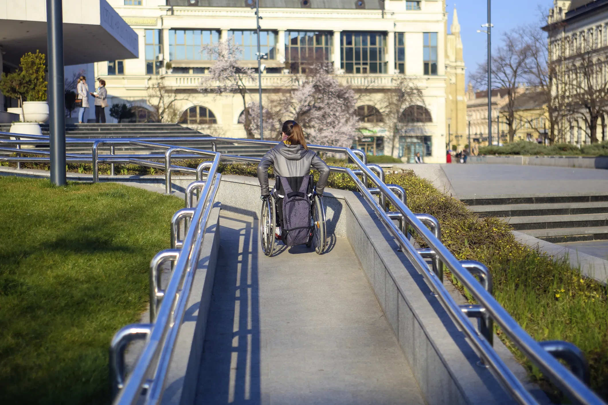Woman in wheelchair moving up ANSI A117.1 ramp with blue railings outdoors.