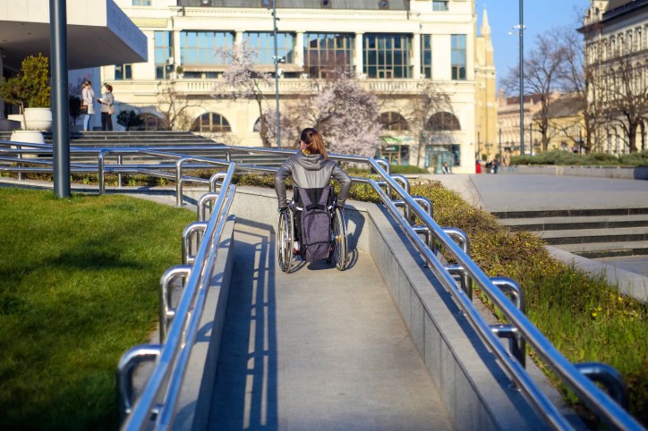 Woman in wheelchair moving up ANSI A117.1 ramp with blue railings outdoors.