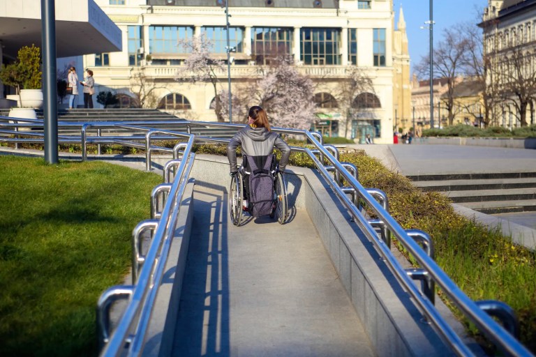 Woman in wheelchair moving up ANSI A117.1 ramp with blue railings outdoors.