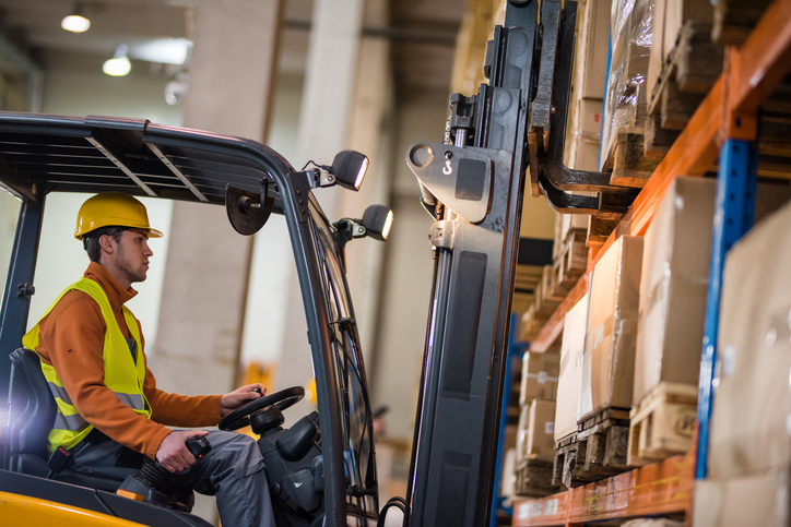 Man properly dressed in yellow and orange PPE to properly operate an ANSI B56.1-2020 forklift on a factory palette.