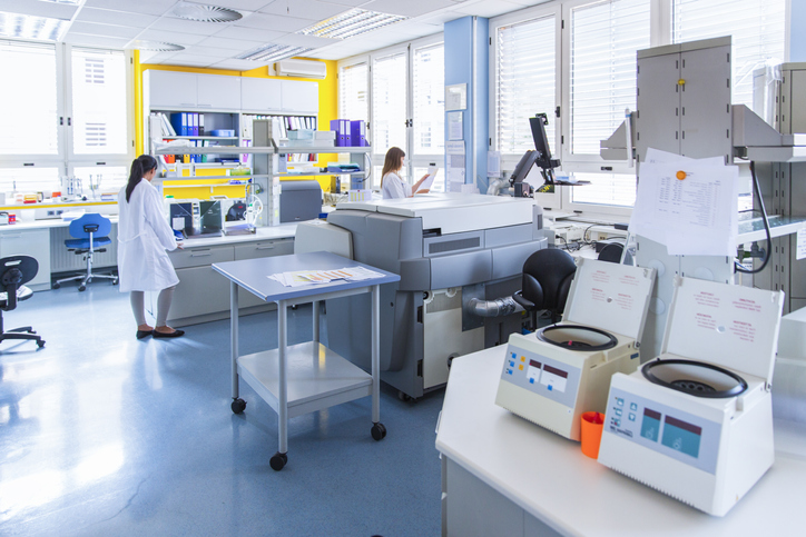 Two female lab technicians in cluttered calibration lab reviewing the ISO/IEC 17025:2017 resource requirements.