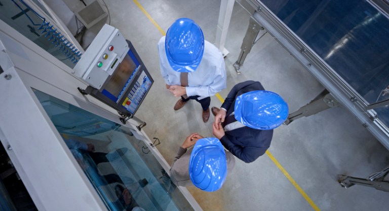 Three managers in blue hardhats abiding to ANSI B11 standards while looking at a safe machine in factory.
