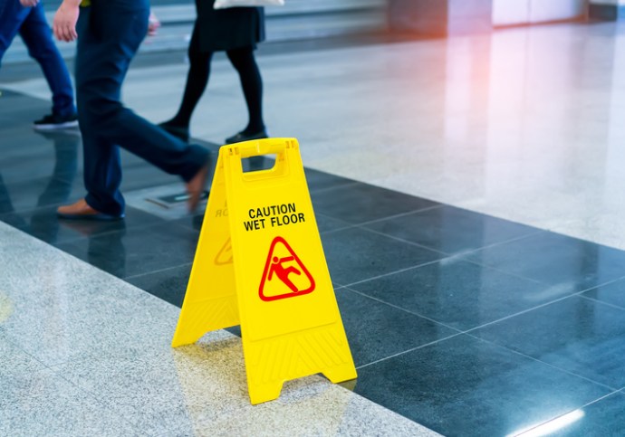 A bright yellow wet floor sign set before walking people on a walkway tested for NFSI B101.1-2022 wet SCOF.