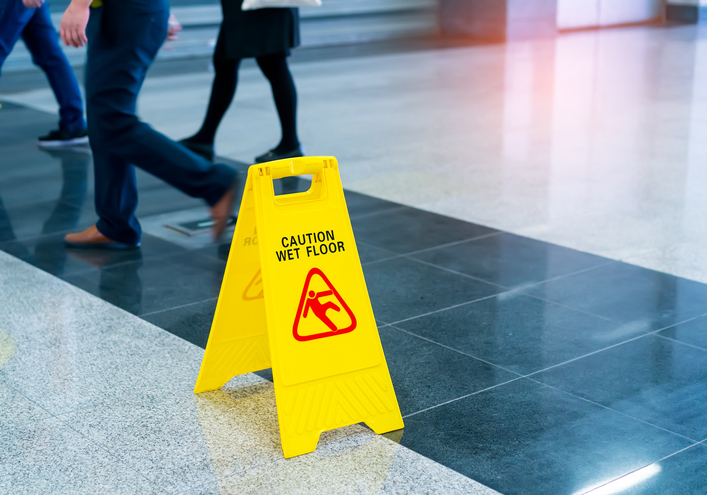A bright yellow wet floor sign set before walking people on a walkway tested for NFSI B101.1-2022 wet SCOF.