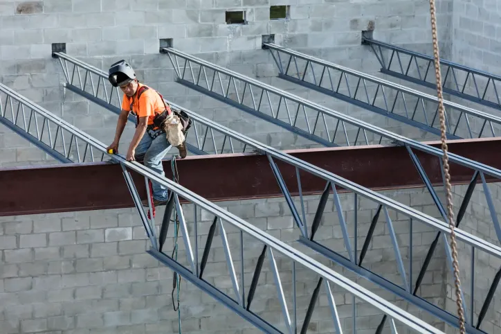 Welder working on metal rafters at height with ANSI/ASSP Z359.1 fall protection requirements.