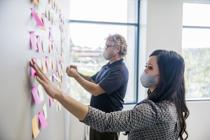 Two project managers wearing face masks and organizing tasks in accordance with ISO 21502:2020 guidance.