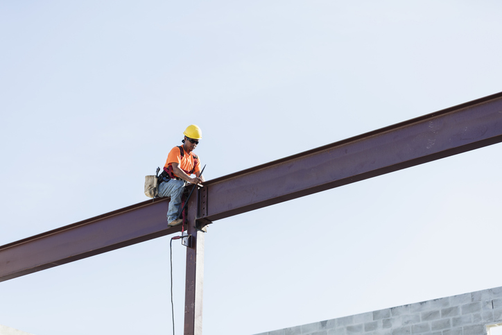 Worker sitting on girder and held safe by ANSI/ASSP Z359.11-2021 full body harness in the sky.