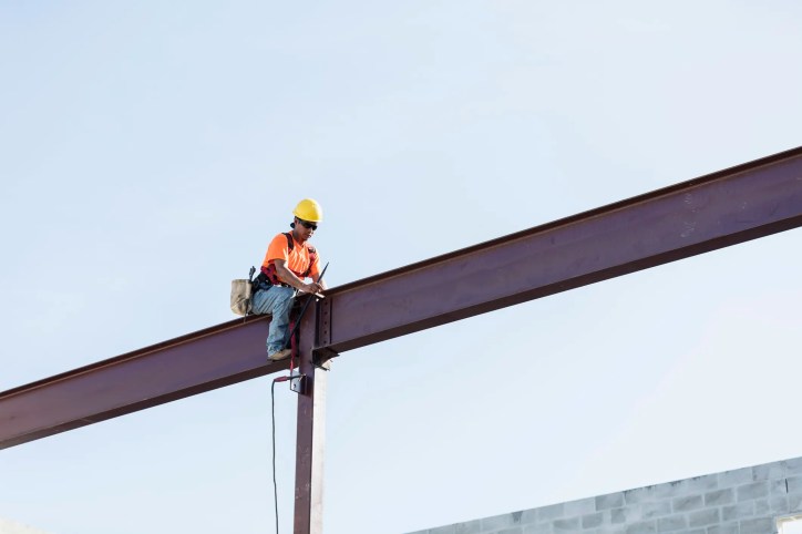 Worker sitting on girder and held safe by ANSI/ASSP Z359.11-2021 full body harness in the sky.