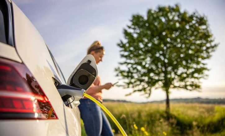 Woman texting while charging electric vehicle in remote area due to growing US charging station infrastructure.