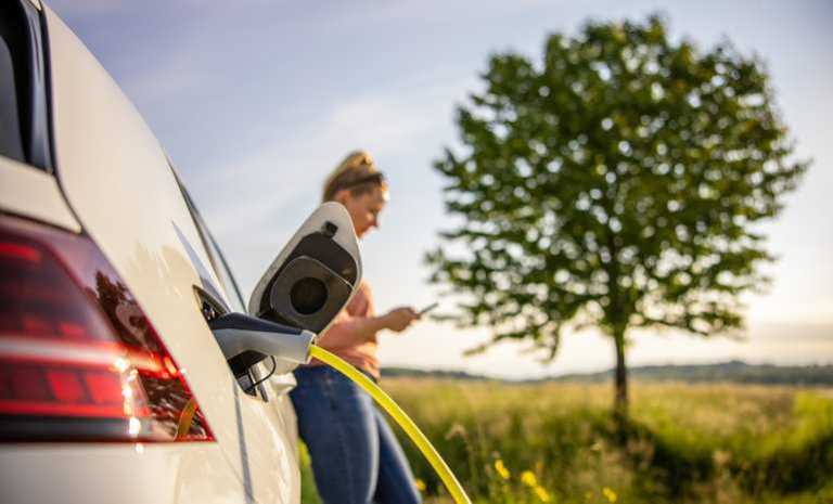 Woman texting while charging electric vehicle in remote area due to growing US charging station infrastructure.