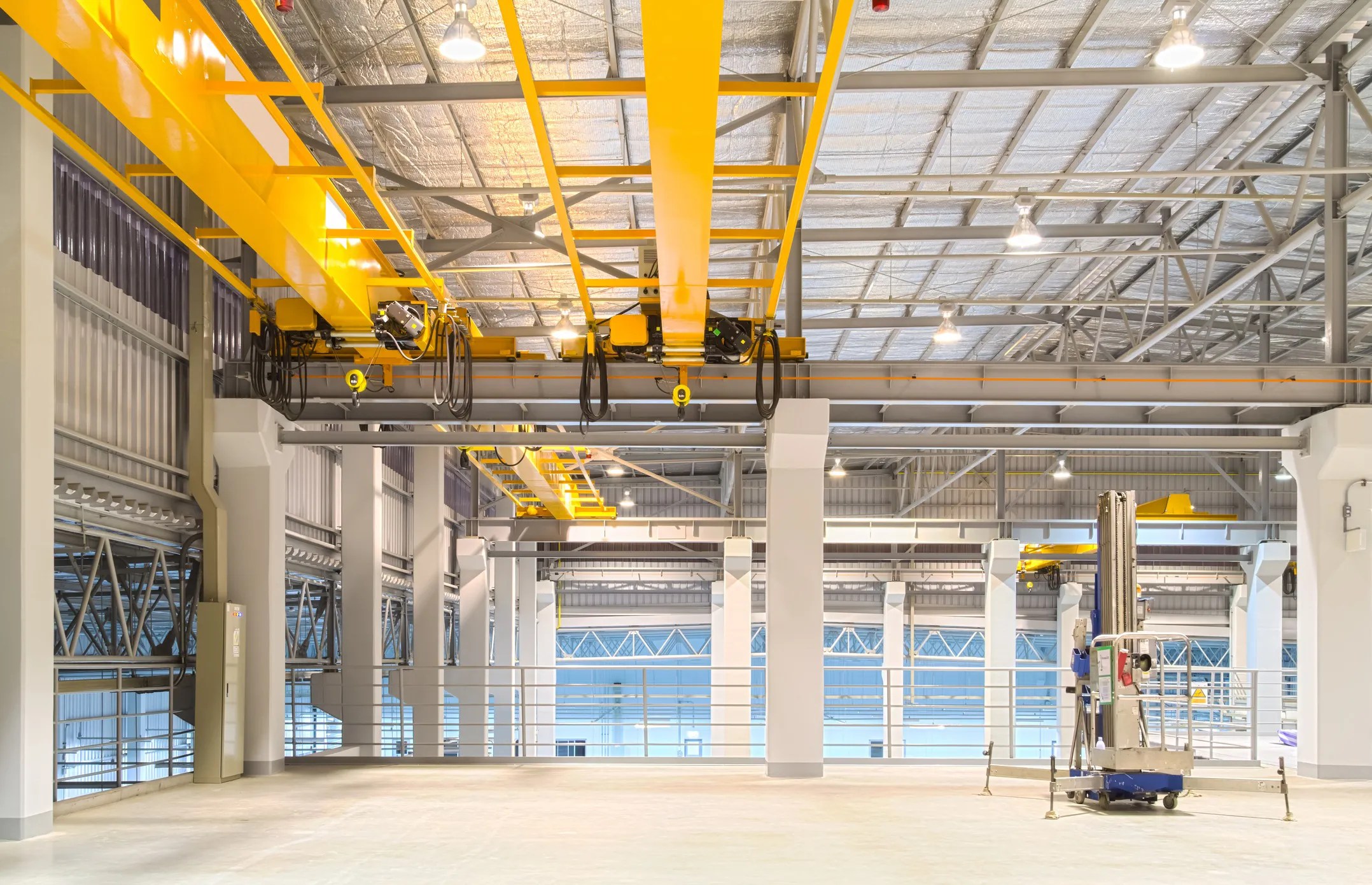A large gantry crane inside a warehouse, featuring a steel frame structure with two vertical supports and a horizontal beam spanning the workspace. The crane’s hoist is suspended from the beam, lifting a heavy pallet of industrial materials.