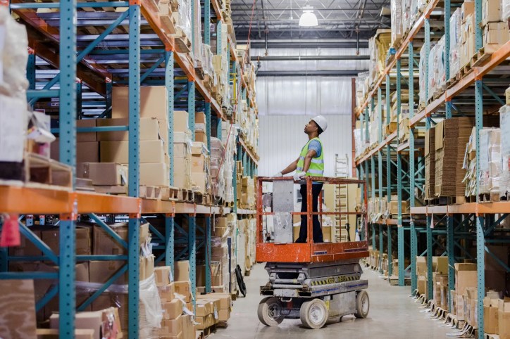 Worker standing on orange scissor lift in warehouse following ANSI MH29.1-2020.