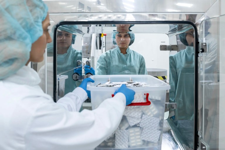 Pharmaceutical workers dressed in full cleanroom attire, including hairnets, face masks, gloves, and protective gowns, carefully operate advanced equipment inside a sterile, brightly lit cleanroom that adheres to ISO 14644-4.