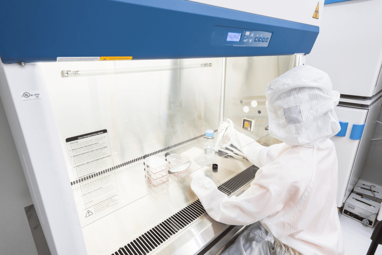 Scientist wearing white full-body PPE while working with pipettes in biosafety cabinet.