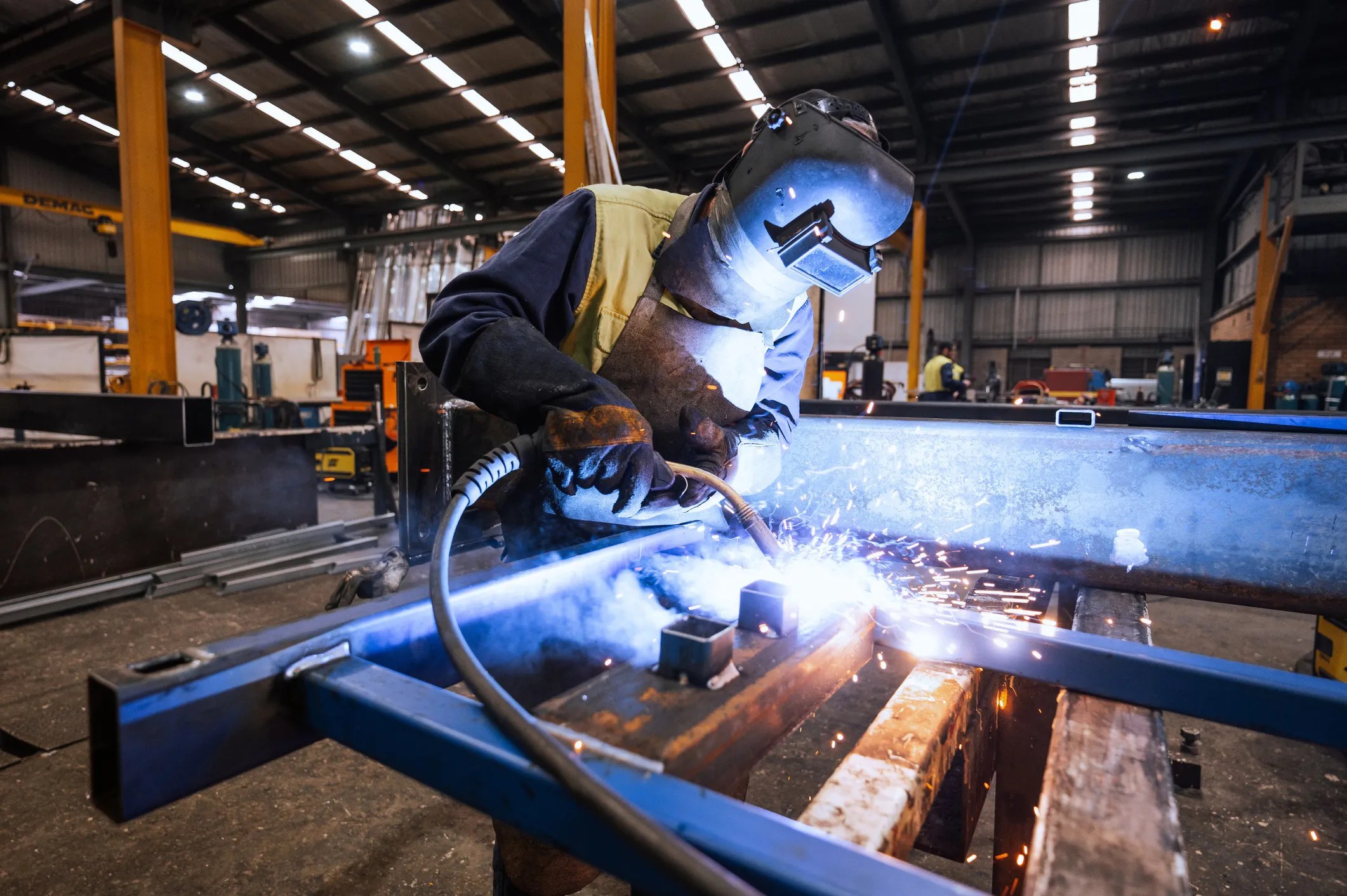 Professional welder adhering to ISO 13920:2023 guidelines and using MIG welding torch on metal frame in industrial workshop.