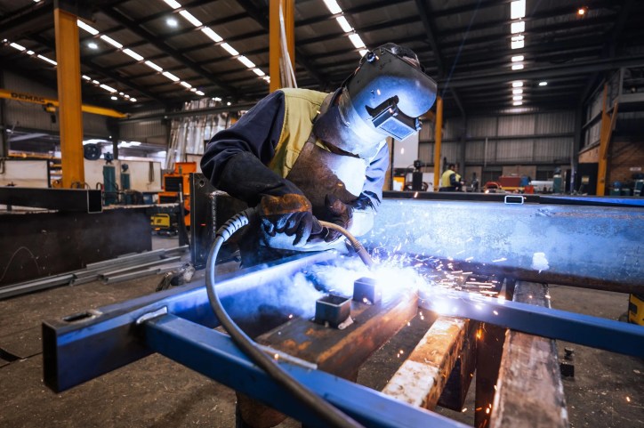 Professional welder adhering to ISO 13920:2023 guidelines and using MIG welding torch on metal frame in industrial workshop.