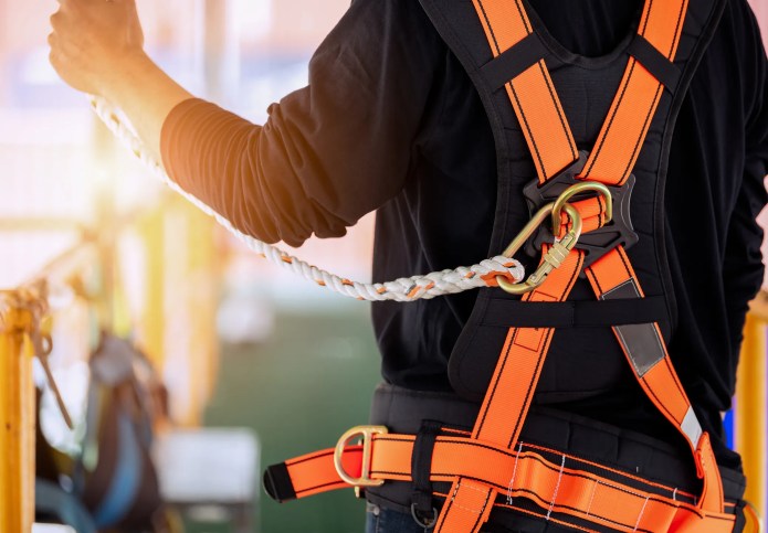 Worker wearing full fall protection gear including a safety harness, lanyard, and helmet while performing tasks at height on a construction site, demonstrating compliance with ANSI/ASSP Z359.2-2023 fall protection standards.
