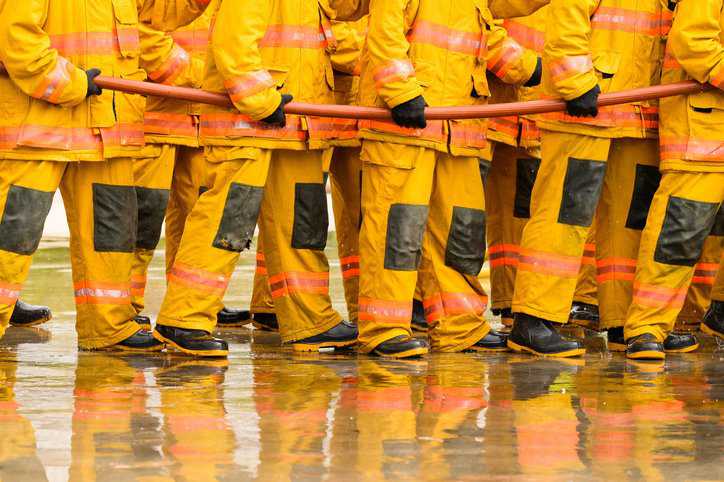Fireman fighting fire flame with water and extinguisher wearing flame-resistant protective clothing that adheres to ISO 11612:2015.