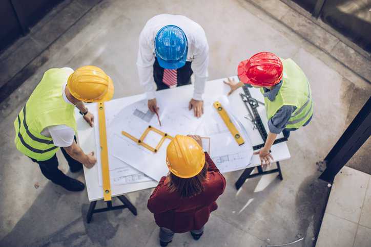 Construction workers and architects working on construction site meeting about pre-project planning, adhering to ANSI/ASSE A10.1-2011 (R2017).