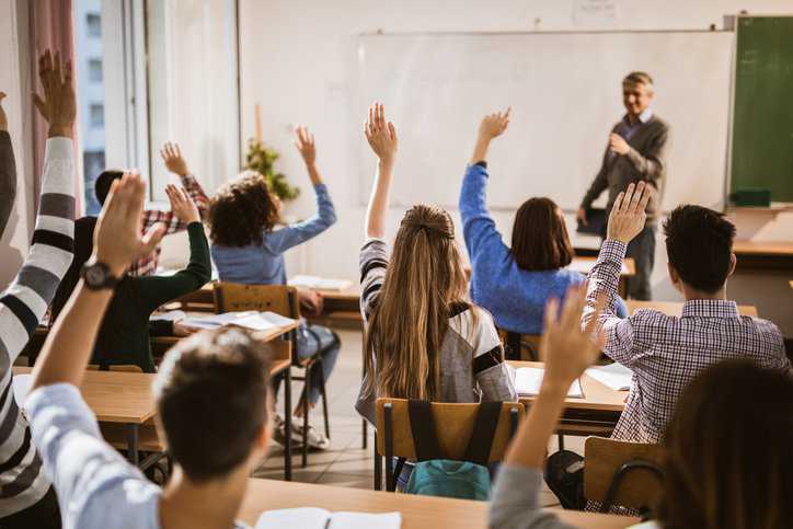 Rear view of large group of students raising their hands to answer the question in classroom with acoustical performance adhering to ANSI/ASA S12.60/Part 1-2010 (R2020).