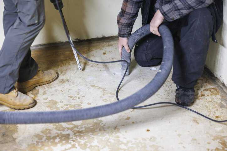 Two construction workers are making a hole in the basement floor to install a radon mitigation system adhering to ANSI/AARST MS-PC-2022.