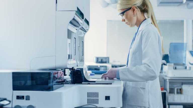 Female lab scientist trained to AR 3125 requirements looking at test tube samples in lab cabinet machine.