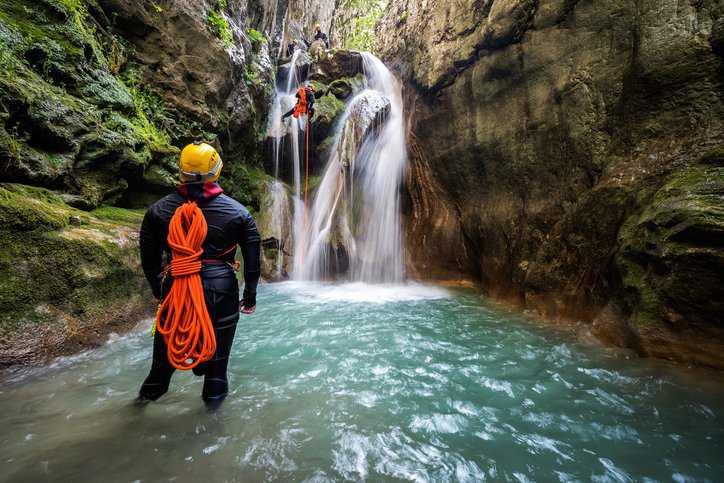 Canyoneering team member with backpack rappeling down the waterfall in the canyon while man in front view and others on top of the waterfall are on stand by with technical rope and harness adhering to ASTM F1772-17.