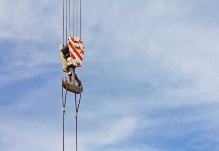 Iron hook ASME BTH-1-2023 below-the-hook-device held by red and white crane over blue sky.