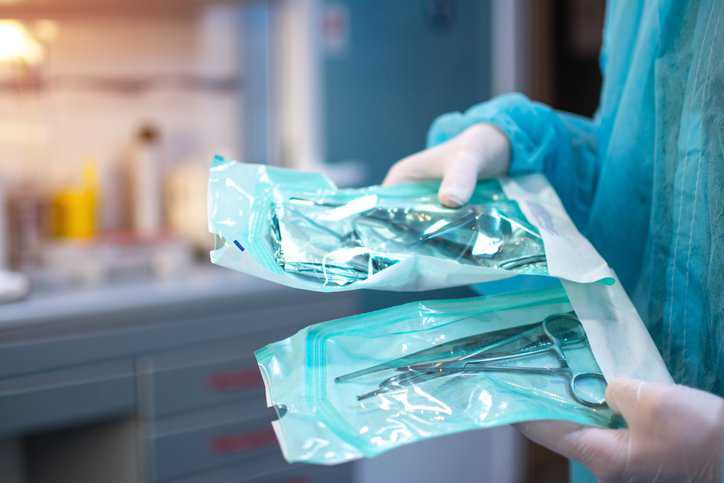 Dentist hands in white sterile gloves holding dental tools for surgical use packed in a protective foil that adheres to ASTM F1929-23 medical packaging requirements.
