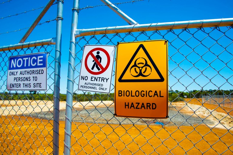 Various ANSI Z535.2‑2023 compliant environmental safety signs outside a hazardous facility. One sign displays a black triangle on a yellow background that states "BIOLOGICAL HAZARD"; another sign states "NO ENTRY" and has a red circle with a diagonal red line on it on a white background; and the last sign states "NOTICE" in white bold letters on a blue background.