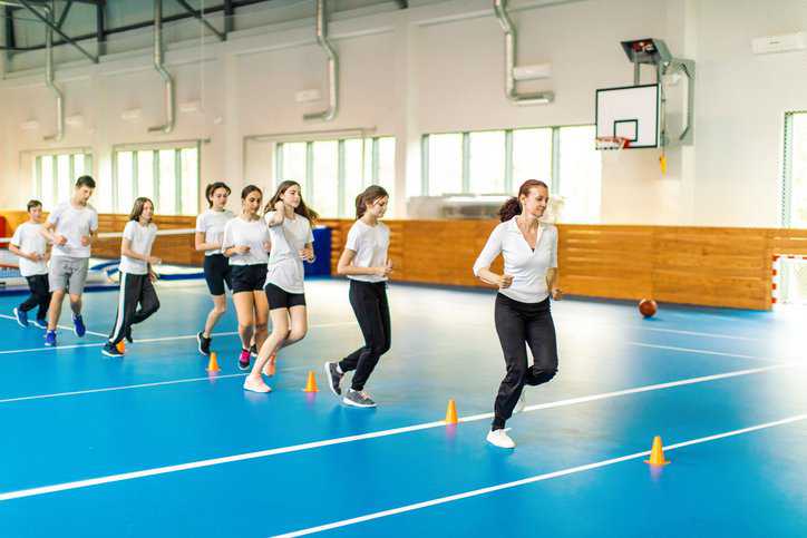 Students and their physical education coach jogging in the school gym that adheres to ASA/ANSI S12.60-2019/Part 4 (R2024) acoustical criteria.