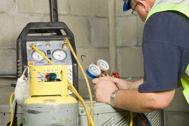 A service technician is using gauges a recovery pump and tank to remove refrigerant from a residential air conditioning system adhering to IEC 60335-2-104:2021.