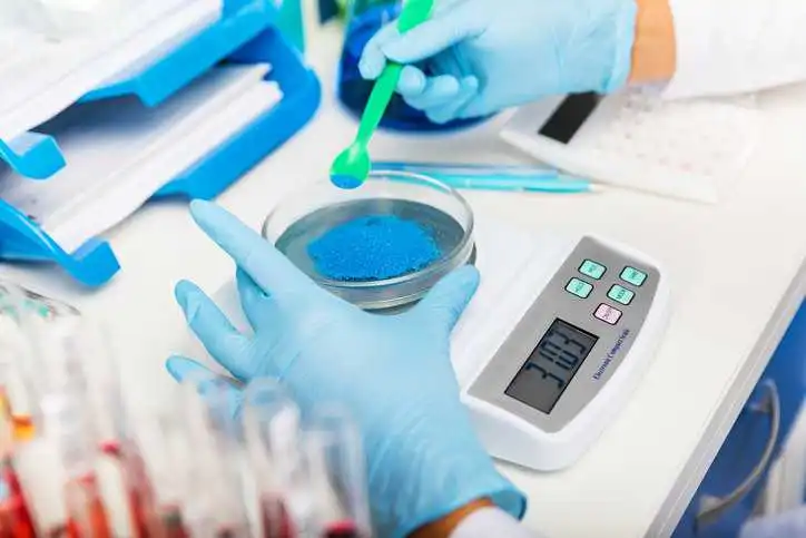 Scientist in gloves measuring weight of blue bulk solid substance in the scientific chemical laboratory assuring ISO 17034 metrological traceability.