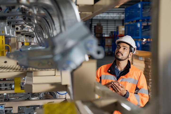Engineer in hardhat reviewing IAF MD on his phone while auditing metal in warehouse.
