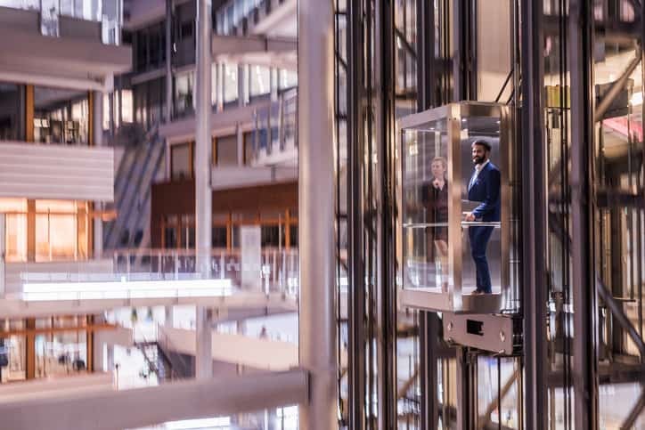 Businesspeople standing in clear existing elevator adhering to ASME A17.3-2023 on way to meeting in fancy, blurry building.