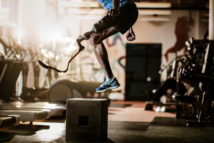 Unrecognizable man with artificial leg jumping on crate during gym training in an inclusive fitness environment that adheres to ANSI/RESNA IF-1:2018.