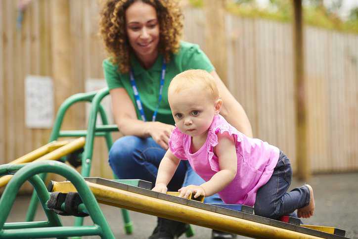 Nursery worker with infant in playground that adheres to ASTM F2373-11(2017).