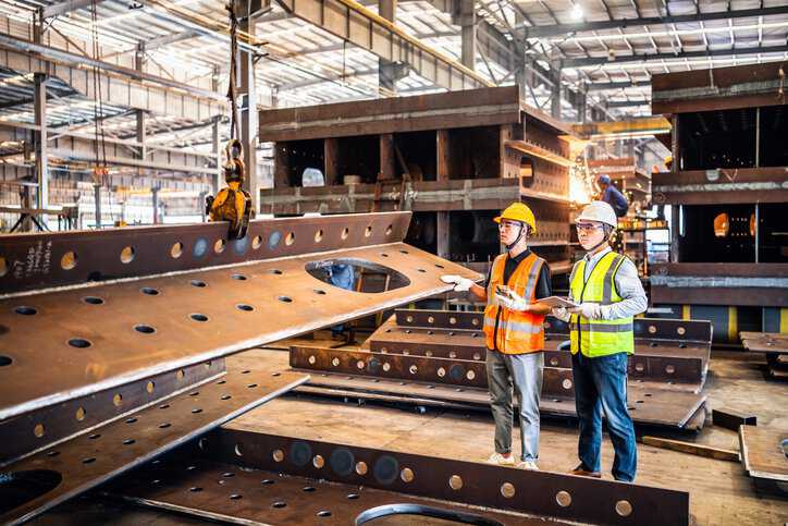 Two metal workers operating a crane to move steel members adhering to machine safety risk assessment processes in ANSI/ISO 12100:2012.