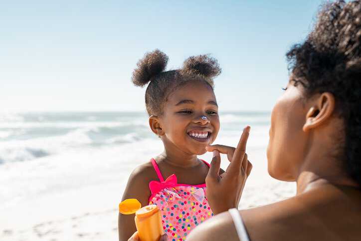 Mother applying protective sunscreen on daughter's nose at beach with sunscreen adhering to SPF specifications in ISO 24442:2022.