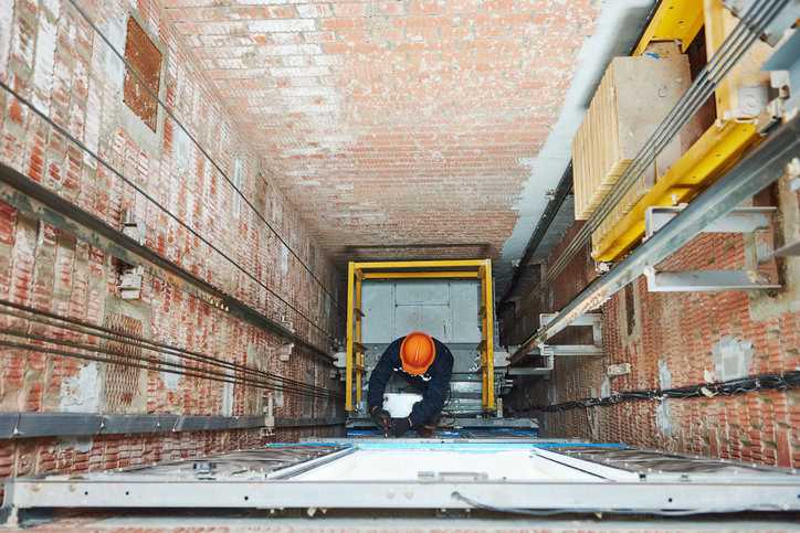 With the ASME A17.3-2023 guide in hand, one professionals in hardhat inspects an elevator in its shaft.